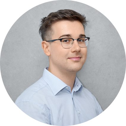 Mladý muž v brýlích a světle modré košili s krátkými vlasy, s mírným úsměvem, na šedém pozadí.