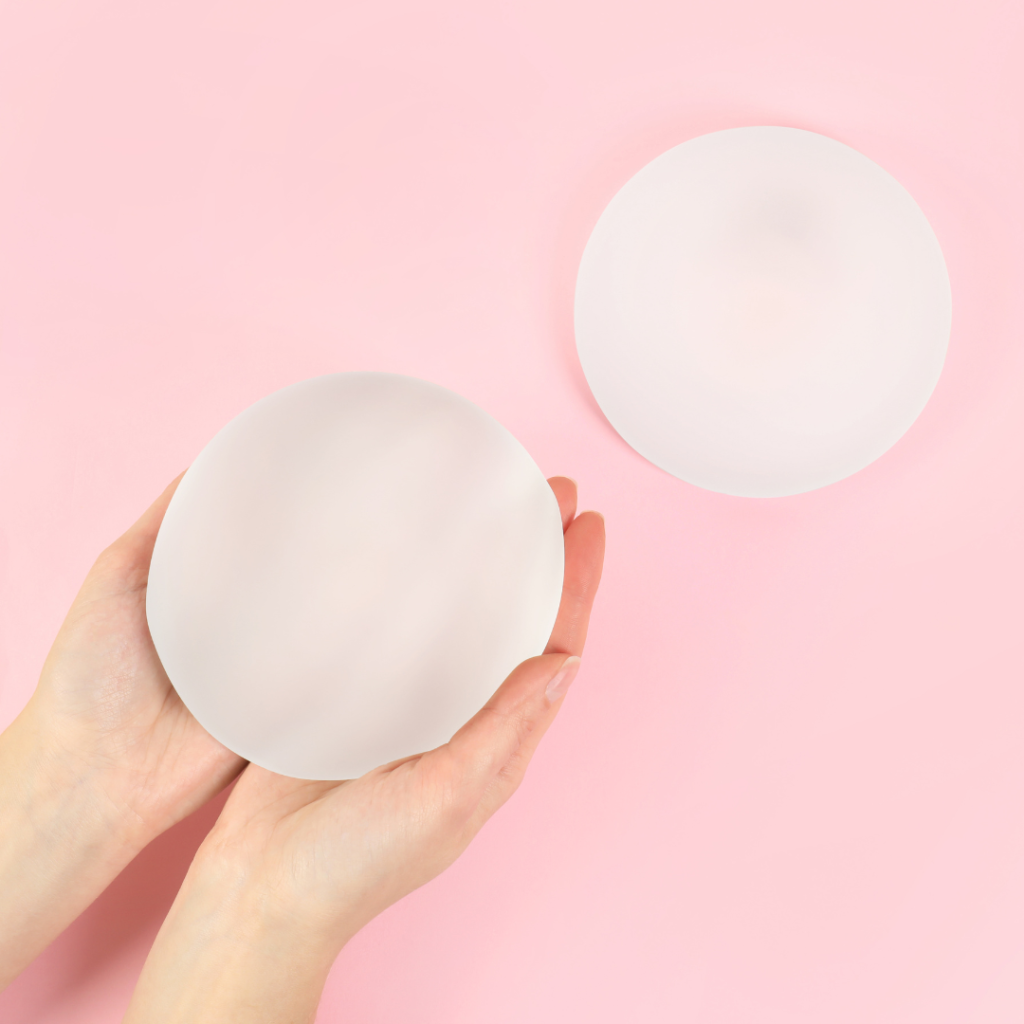 Dva silikonové prsní implantáty na růžovém pozadí, jeden držený ženskýma rukama, druhý volně položený.